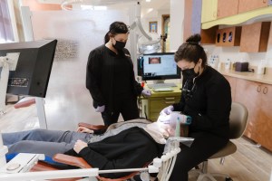 Two dental assistants wearing masks and scrubs work together on a patient in a clinical treatment chair.