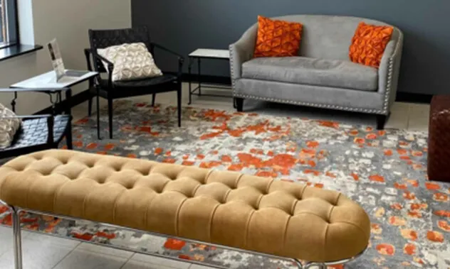 A modern, cozy waiting room with a grey sofa, orange pillows, a tufted bench, and a decorative patterned rug.