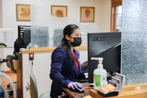 A staff member wearing a face mask works at a computer behind a glass partition at a dental office front desk.