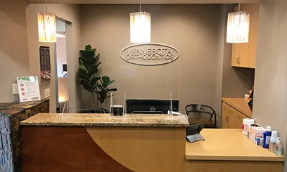 A warm and professional reception area with a curved wooden desk, hanging pendant lights, and the Minnesota Orthodontics logo on the back wall.