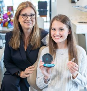 A female dentist stands next to a smiling young woman who is holding a blue dental aligner case.