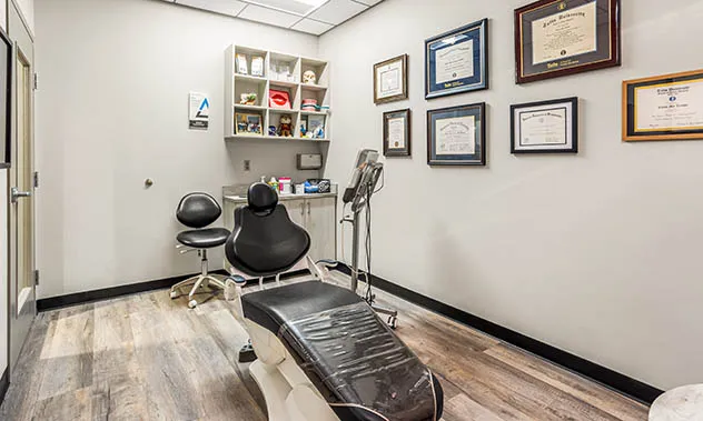 A professional consultation room with a black patient chair and a wall covered in framed certificates and diplomas.