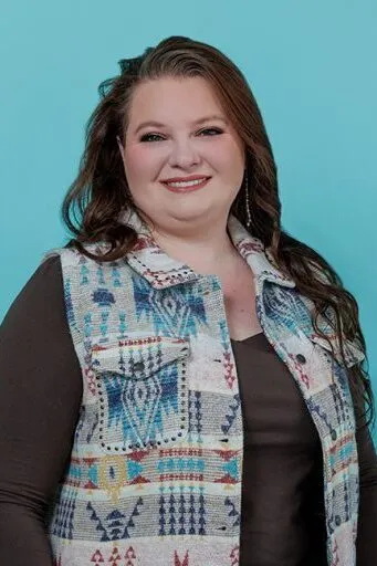 A headshot of Kayla M. – Treatment Coordinator with dark hair wearing a patterned blue and white top against a light blue background.