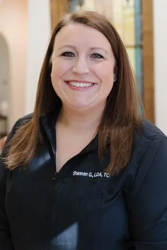 A headshot of Shannan G. – Treatment Coordinatorwith dark hair wearing a black professional scrub top.
